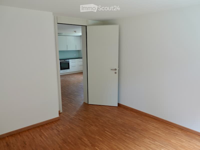 Empty room with wooden flooring and white walls, featuring an open doorway leading to a modern kitchen area with white cabinets and stainless steel appliances.