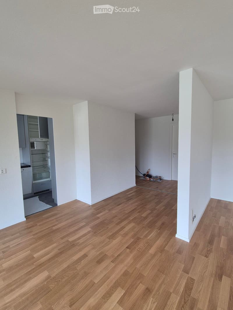 Empty room with wooden flooring and white walls, featuring an open doorway to another room with a refrigerator and a vacuum cleaner visible in the adjacent space.
