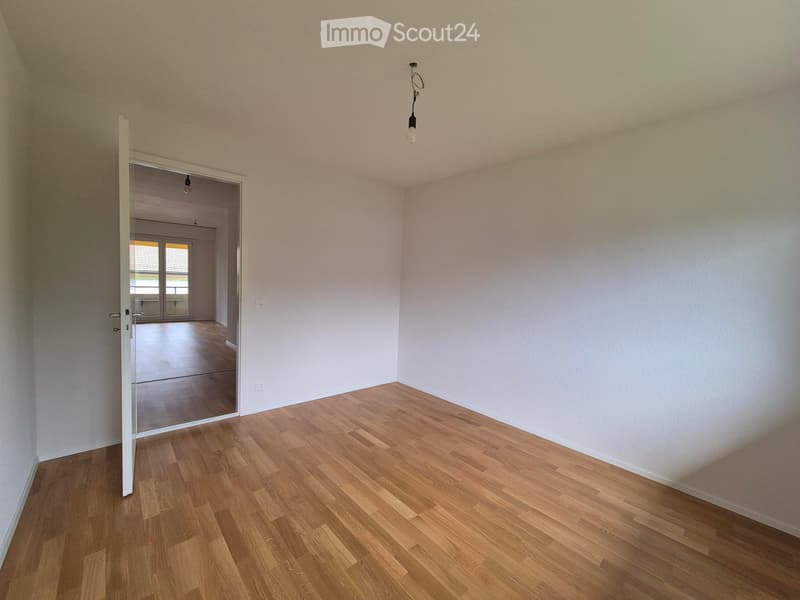 Empty room with light wooden flooring and white walls, featuring a single hanging light fixture and an open doorway leading to another room.