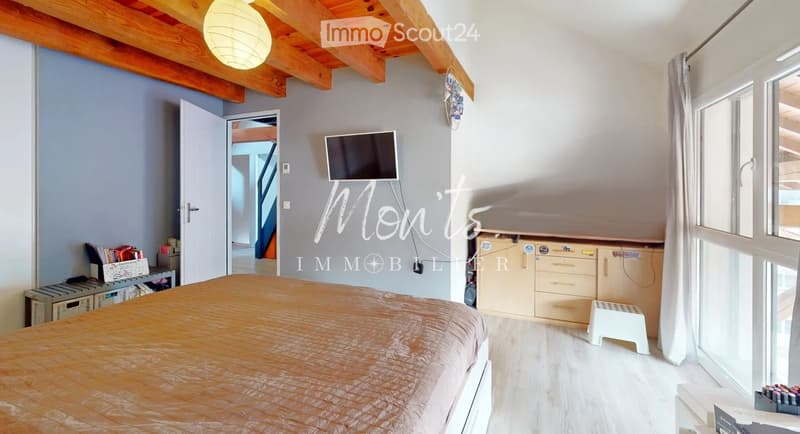 Bright bedroom with wooden ceiling beams, large windows, a bed with a patterned duvet, a nightstand, a television mounted on the wall, a small cabinet, and a white stool.