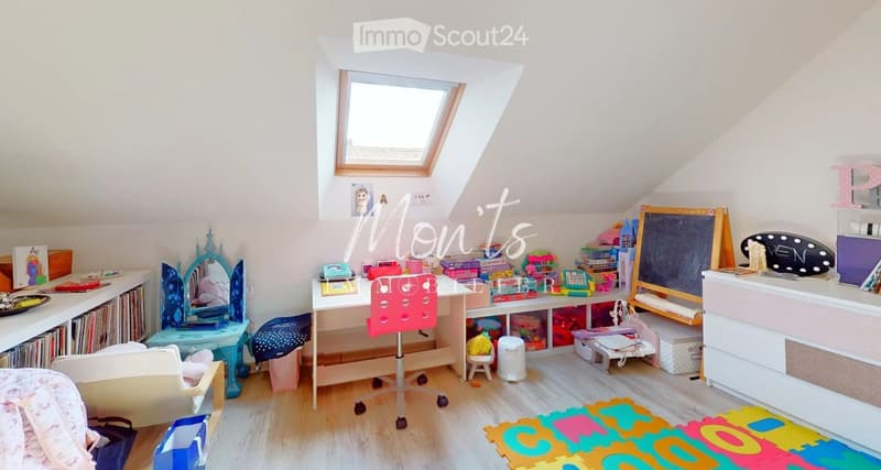 Brightly lit children's room with slanted ceilings, wooden flooring, and a skylight window. Features a desk with a chair, a mirror, shelves filled with toys and books, a small stool, and a colorful play mat on the floor.