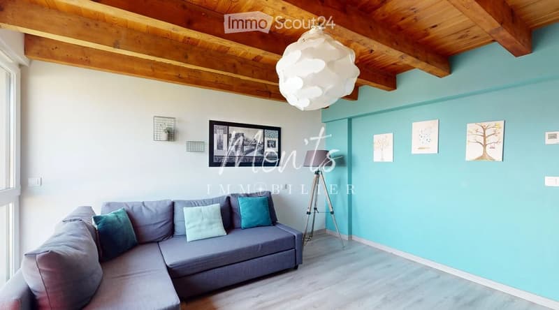 Modern living room with a grey sectional sofa, decorative pillows, a floor lamp, and artwork on the walls. The room features exposed wooden beams on the ceiling and a light blue accent wall.