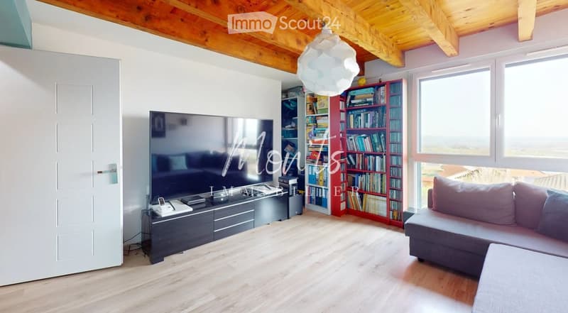 Modern living room with wooden flooring, exposed wooden beams, large windows, grey sofa, flat-screen TV, bookshelf, and a hanging lamp.