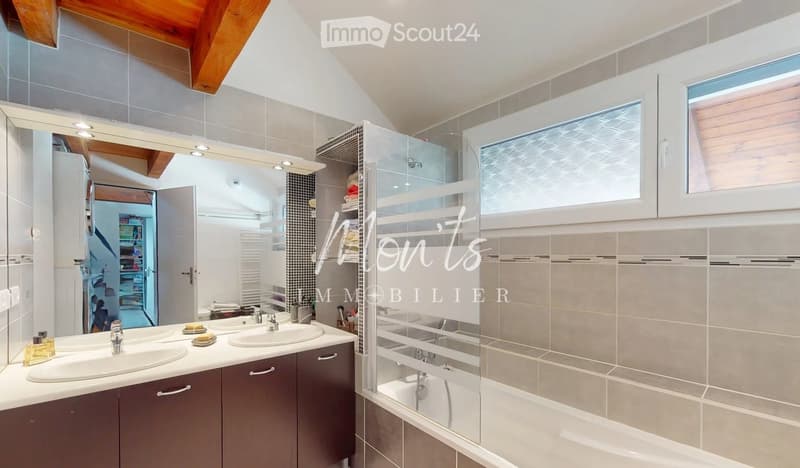 Modern bathroom with double sink vanity, large mirror, bathtub, and built-in cabinets. Features a sleek design with grey tiles and wooden accents.