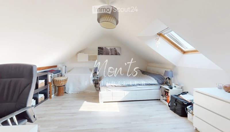 Bright attic bedroom with wooden flooring, slanted ceilings, and skylight windows. Features a white bed, a black chair, a small table with a lamp, a wooden stool, and a nightstand with books and a suitcase.