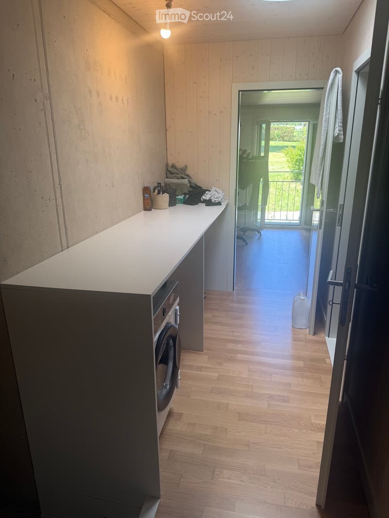 Hallway with wooden flooring, white walls, and a small counter with a washing machine. A door leads to a balcony area with outdoor view.