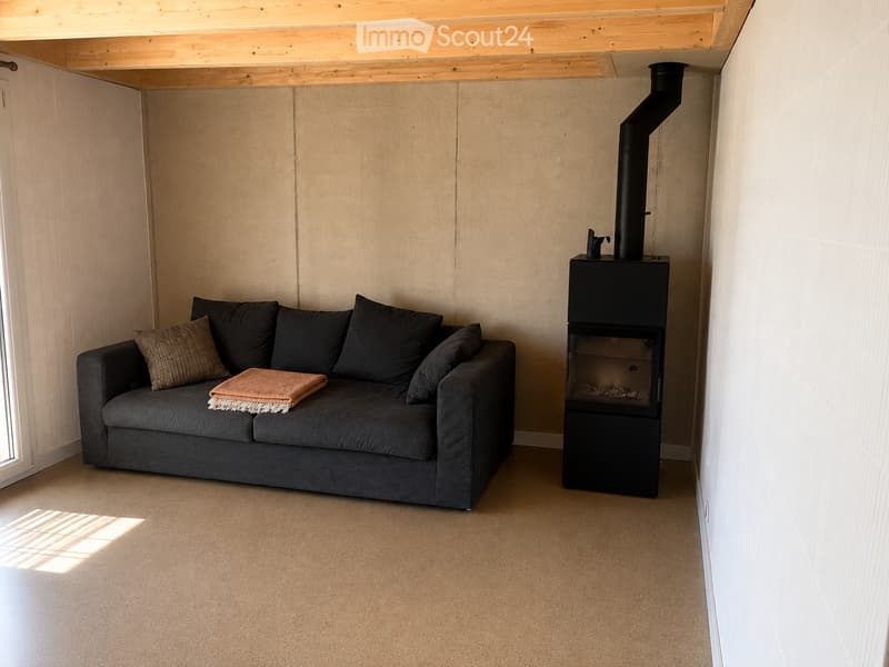 Modern living room with a grey sofa, throw blanket, and a black wood-burning stove with a chimney. The room features light-colored walls and a wooden ceiling.