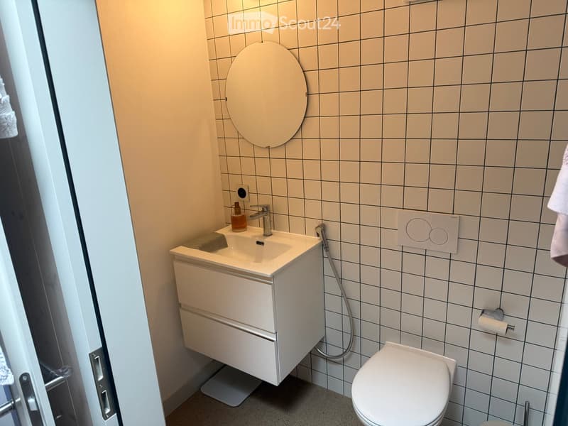 Modern bathroom with white tiled walls, a white sink with a chrome faucet, a toilet, and a round mirror above the sink.