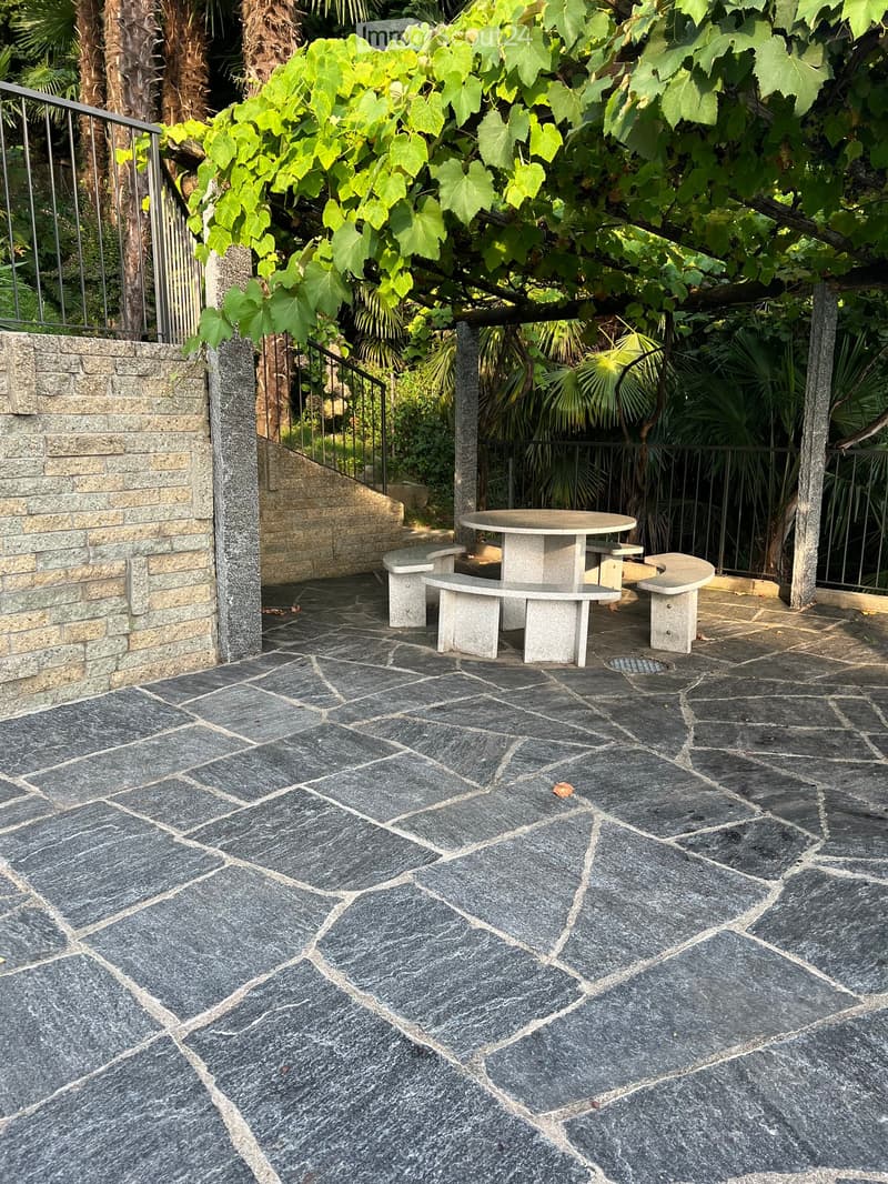 Outdoor patio with stone tiles, a round stone table, and benches surrounded by lush greenery and a metal railing.