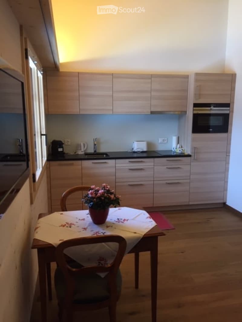 Modern kitchen with light wooden cabinets, black countertops, and a small dining area with a table and chairs. A vase with flowers is placed on the table.
