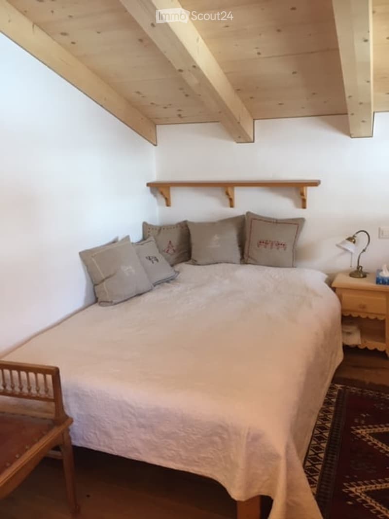 Cozy attic bedroom with wooden slanted ceilings, a neatly made bed with multiple pillows, a nightstand with a lamp, a wooden shelf, and a small wooden chair.