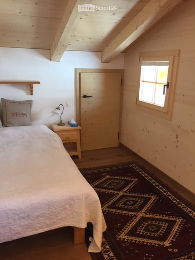 Cozy bedroom with wooden beams, a bed with white bedding, a nightstand with a lamp, a patterned rug, and a wooden door.