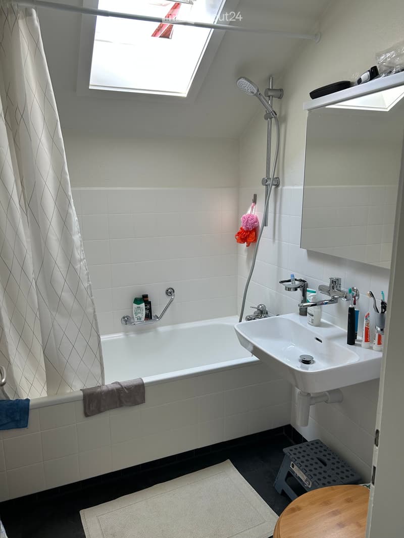 Modern bathroom with a bathtub, sink, mirror, and shower. Features a window with a curtain, a towel on the edge of the tub, and a small wooden stool near the sink.