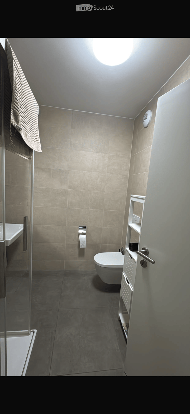 Modern bathroom with beige tiles, a toilet, a sink, a mirror, a small cabinet, and a towel hanging on the wall.