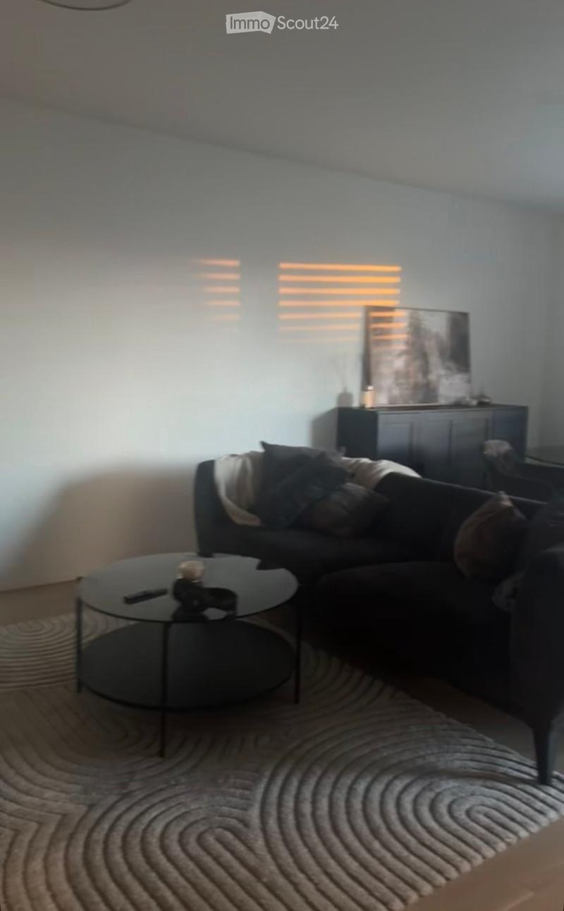 Modern living room with a dark grey sectional sofa, a round glass coffee table, a patterned rug, and a wall-mounted art piece.