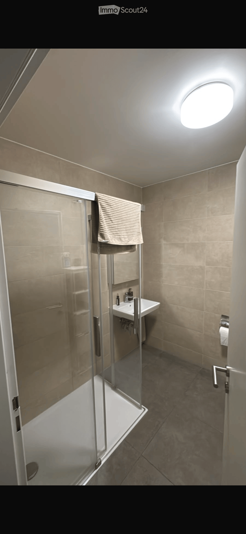 Modern bathroom with a glass-enclosed shower, a white sink with a chrome faucet, and a towel hanging on a rack. The walls are tiled in a warm beige color, and the ceiling features a round skylight.