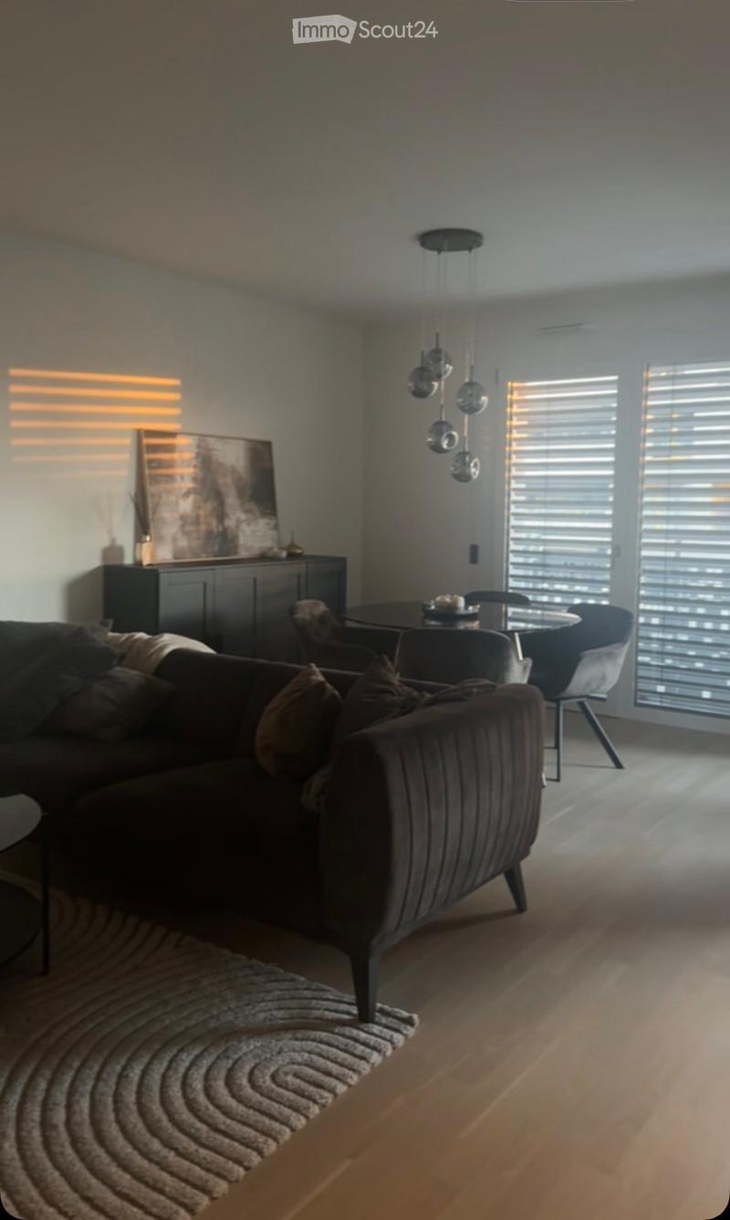 Modern living room with a dark brown sofa, a round dining table with chairs, a cabinet with artwork, pendant lights, and a patterned rug on the wooden floor.