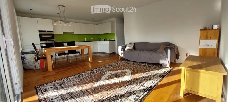 Open-plan living and dining area with a grey sofa, coffee table, dining table with chairs, and a modern kitchen with white cabinets and stainless steel appliances.