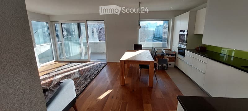 Bright, open-plan living and dining area with wooden flooring, large windows, a sofa, dining table, chairs, and a modern kitchen with white cabinets and an oven.