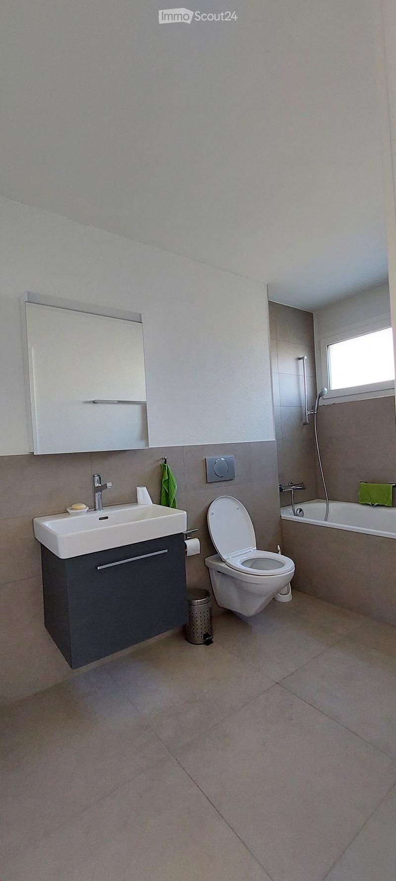 Modern bathroom with a white sink, toilet, bathtub, and mirror. Features a dark grey cabinet under the sink and a small window above the bathtub.