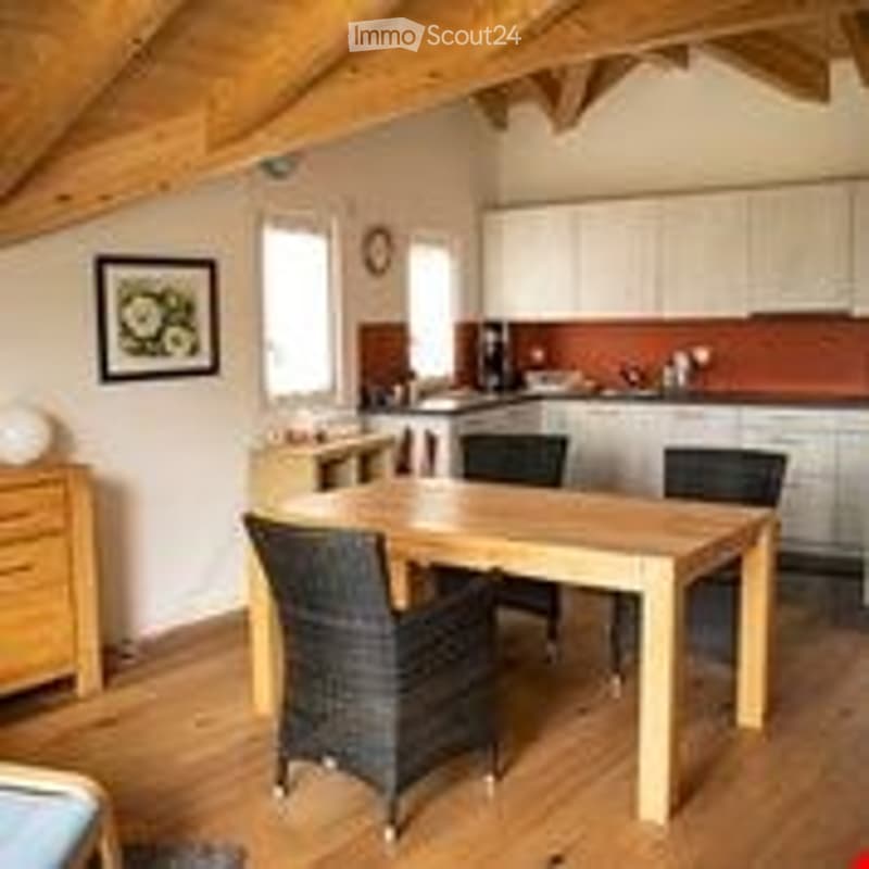 Bright, airy dining area with exposed wooden beams, a wooden dining table with four black chairs, white kitchen cabinets, and a small nightstand with a picture frame on it.