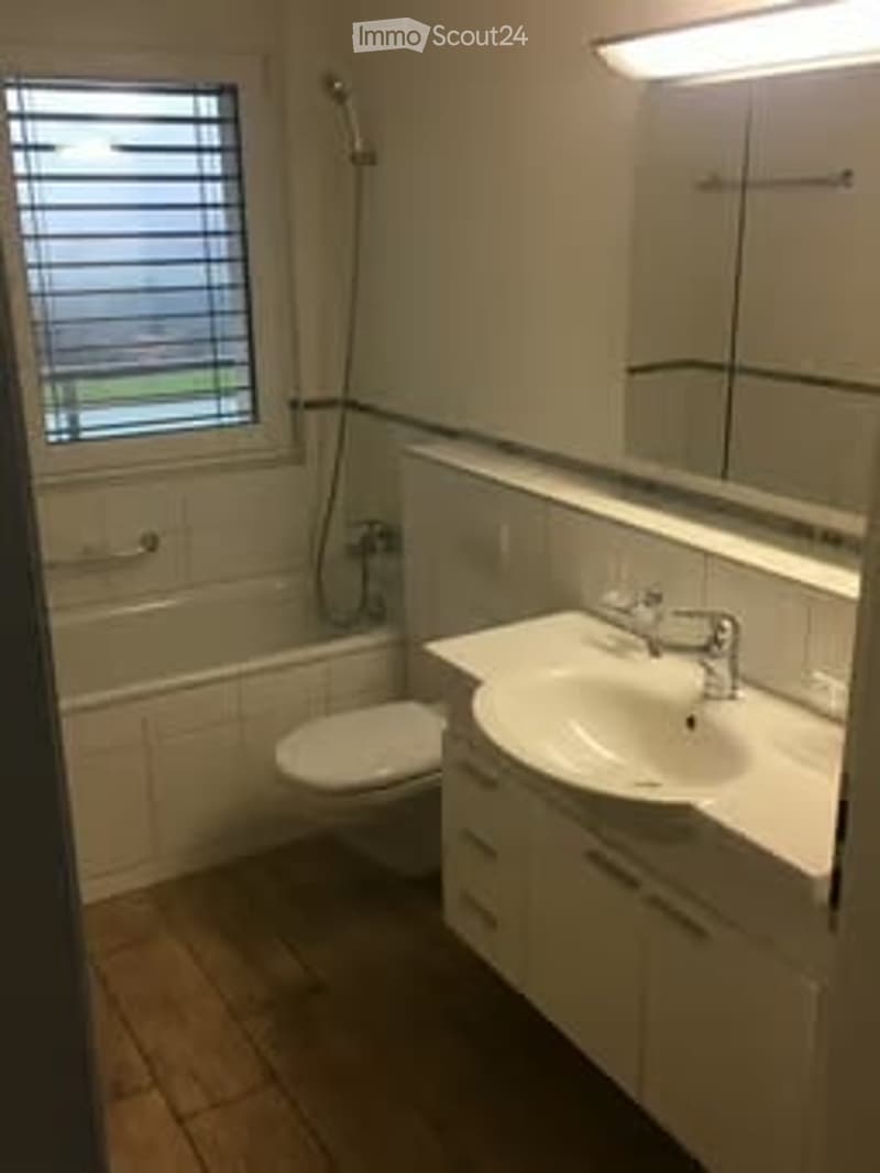 Modern bathroom with a bathtub, toilet, sink, and mirror. The room features white tiles, a window with blinds, and a wooden floor.