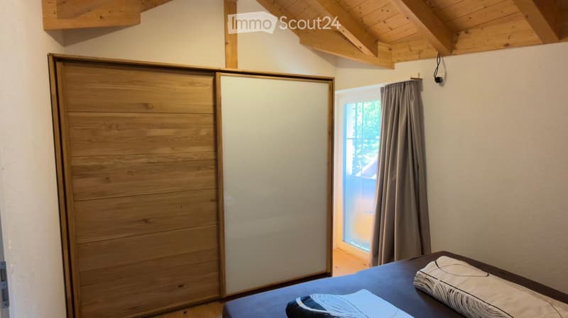 Cozy bedroom with wooden slatted wardrobe, bed with pillows, and a window with curtains.
