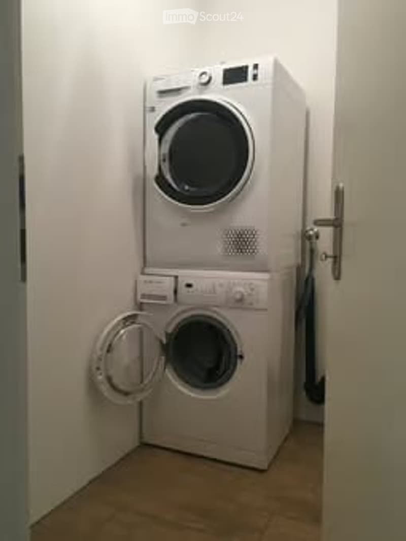 A compact bathroom with a stacked washing machine and dryer unit, white walls, and a wooden floor.