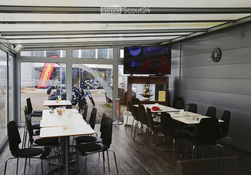 Modern restaurant interior with wooden tables, black chairs, a television screen, and a flower vase on a wooden stand.