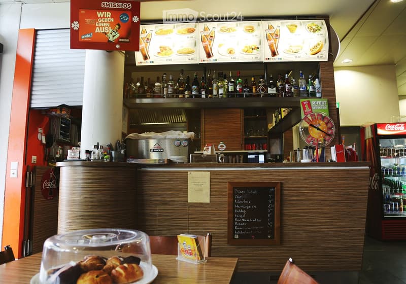 The image shows a restaurant interior with a wooden table displaying a cake, a menu board, shelves with bottles, a refrigerator, and a counter with various items.