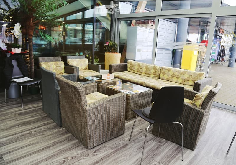 A modern, well-lit living area with wicker furniture, including a sofa, chairs, and a coffee table, arranged on a wooden floor with large windows offering a view of the outside.