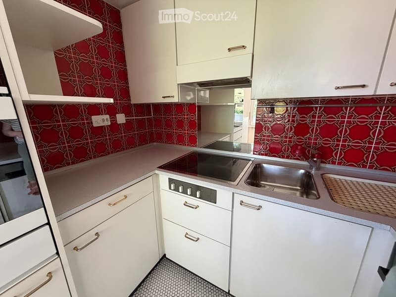 Modern kitchen with white cabinets, stainless steel sink, and electric stove. Features red patterned tiles on the backsplash and a light grey countertop.