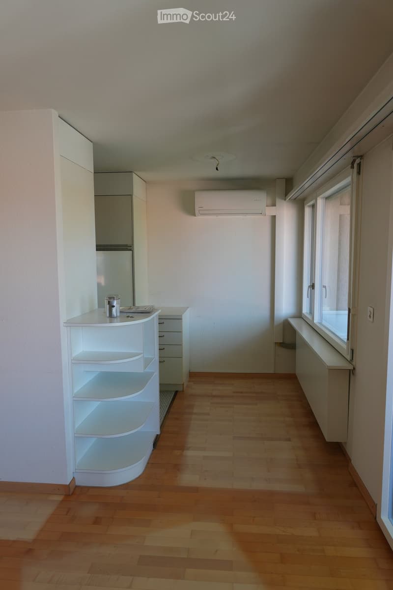 A small, unfurnished room with wooden flooring, white walls, and a window. A white shelf with a cup and remote control is placed near the door.
