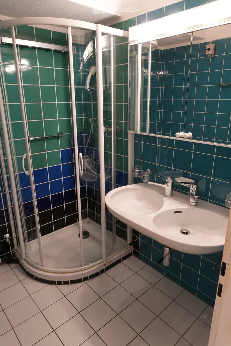 Modern bathroom with white tiled floor, blue and green tiled walls, a corner shower with glass enclosure, a white sink with dual faucets, and a mirror with built-in lighting.