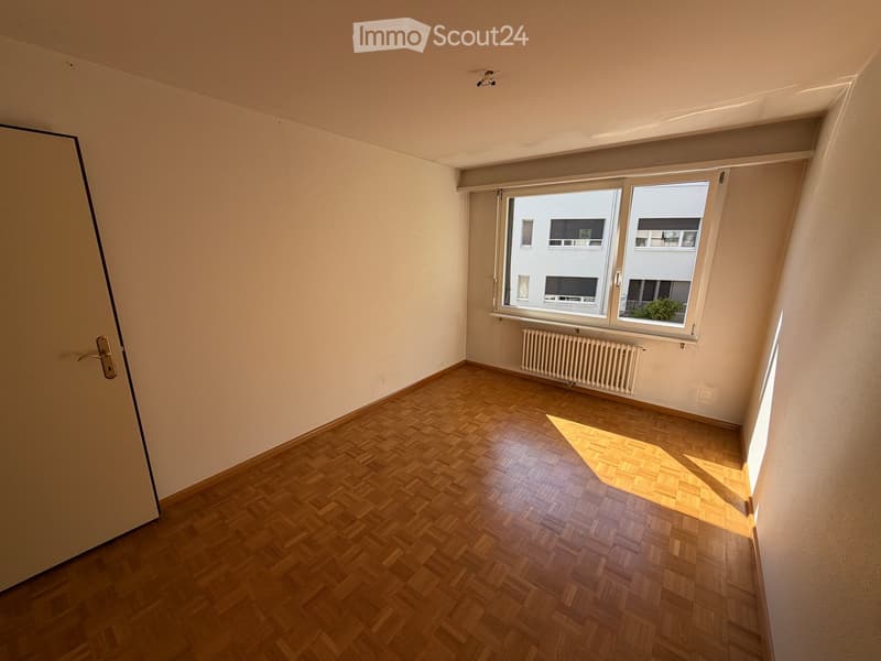 Empty room with wooden herringbone flooring, white walls, and large windows with white frames. A radiator is visible beneath the windows, and a door is on the left side.