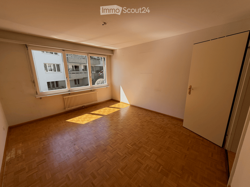 Empty room with wooden flooring, large windows, radiator, and a door leading to another space.