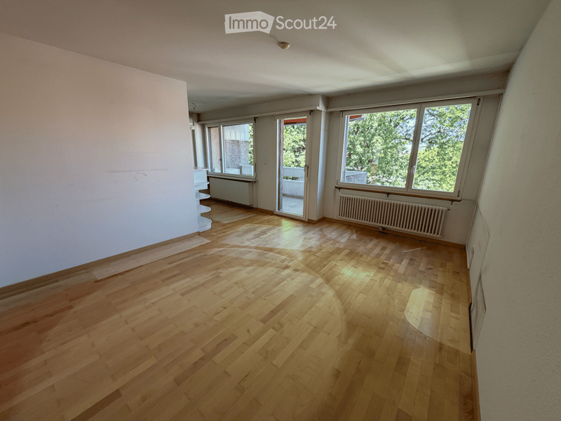 Empty room with wooden flooring, large windows, and a sliding glass door leading to a balcony. The walls are painted in neutral tones, and there are radiators visible along the walls.
