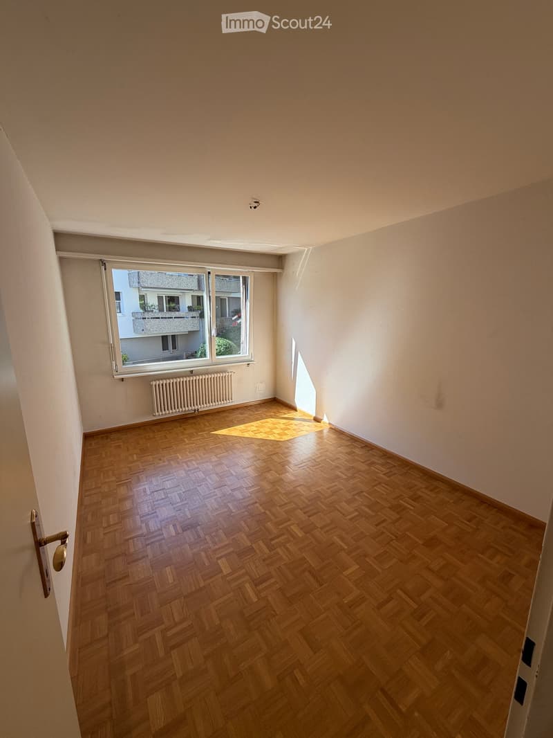 Empty room with wooden parquet flooring, white walls, and a large window with a view of a neighboring building and balcony. A radiator is visible below the window, and a small rug is placed on the floor near the window.