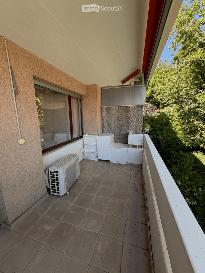 Balcony with tiled floor, white cabinets, and an air conditioner unit, overlooking greenery.