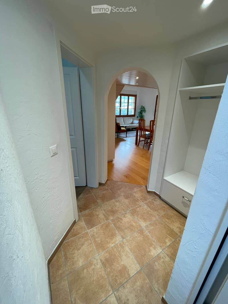 A well-lit hallway with tiled flooring, white walls, and an arched opening leading to a dining area with wooden furniture and a plant.