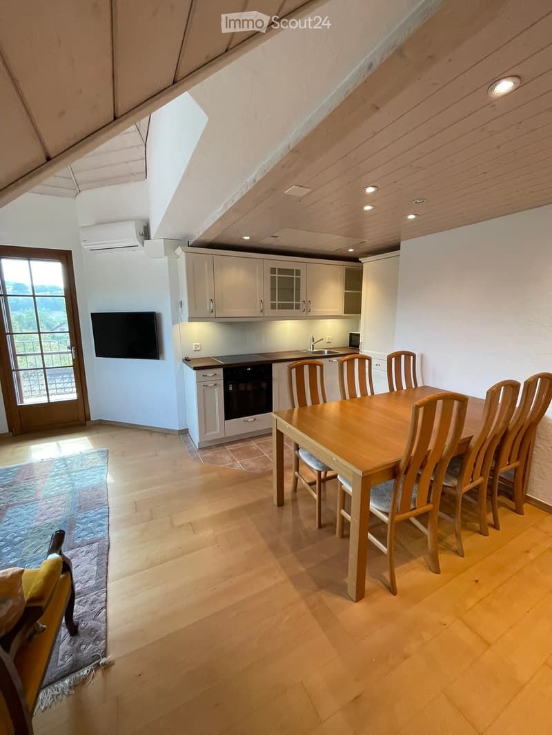 Open-plan kitchen and dining area with wooden flooring, white cabinets, a dining table with six chairs, and a screen mounted on the wall.