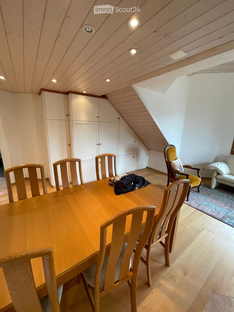 Bright attic room with wooden table, chairs, sofa, and built-in white cabinets under a slanted wooden ceiling with recessed lighting.