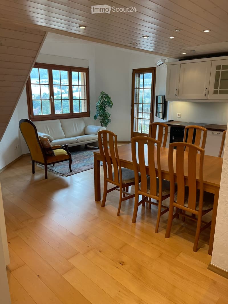 Bright, open living and dining area with wooden flooring, white walls, and large windows. Features a cream sofa, armchair, dining table with chairs, and a small kitchen area with cabinets.