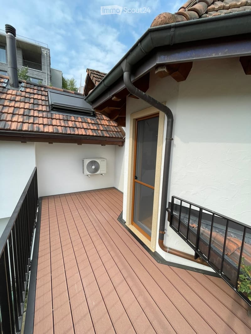Wooden deck with black metal railing, air conditioning unit mounted on wall, adjacent building with tiled roof visible.