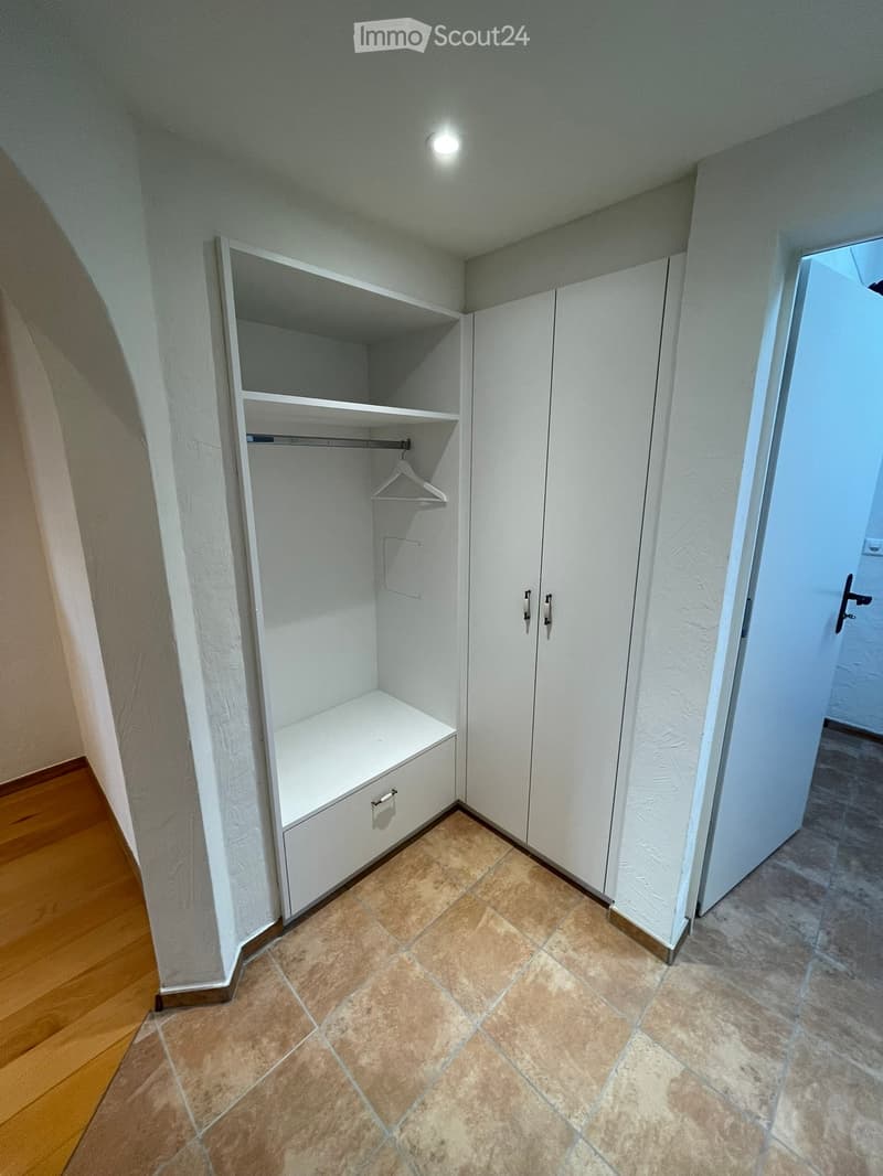 Empty hallway with tiled floor, white walls, built-in closet with shelves and drawers, and a coat hanger. The hallway has a wooden floor on one side and a tiled floor on the other.
