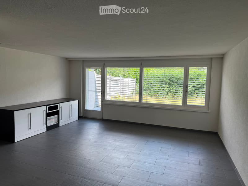 Modern, unfurnished room with dark wooden flooring, white walls, large windows with blinds, and a built-in cabinet with an oven.