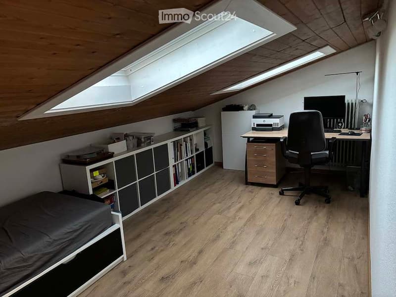 A multi-functional room with a bed, desk, chair, bookshelf, and printer. The room features wooden flooring and slanted wooden ceilings with skylights.