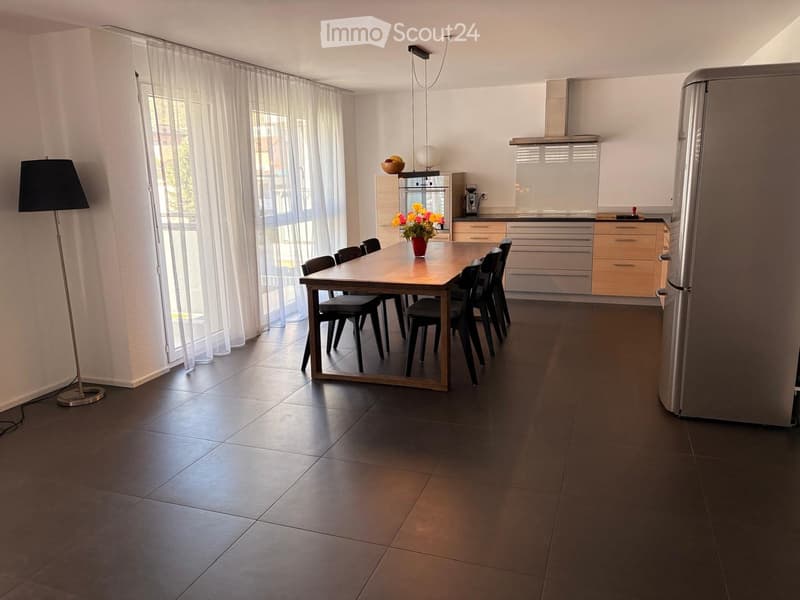 Modern kitchen with a dining area, featuring a wooden dining table, black chairs, a stainless steel refrigerator, and a built-in oven. The space includes a floor lamp and a vase with flowers on the table.