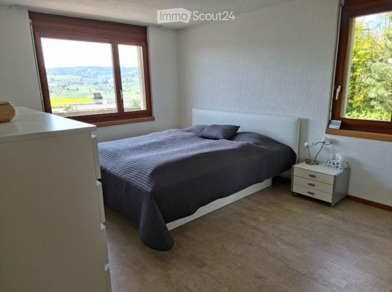 Cozy bedroom with a large window offering a scenic view, a neatly made bed with pillows and a blanket, a nightstand with a lamp, and a drawer unit.