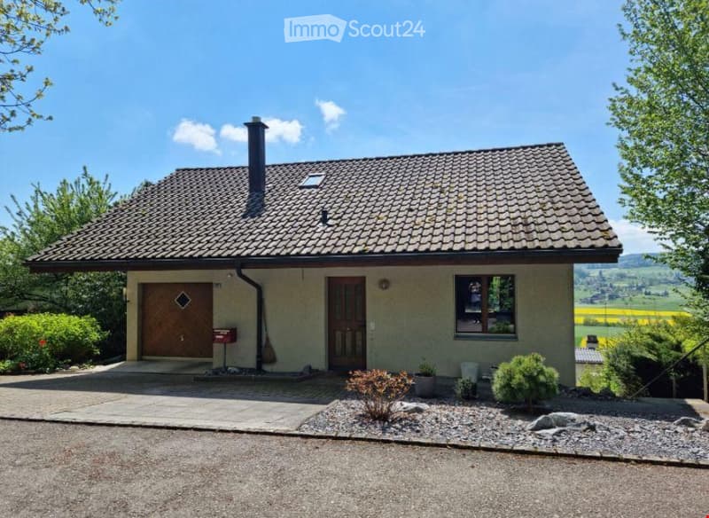 Single-story house with a tiled roof, beige exterior, wooden front door, and small garden area with potted plants.